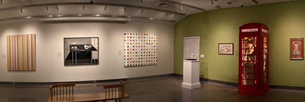 Panoramic photograph of a gallery at Hill‑Stead Museum. Paintings by modern and contemporary artists hang on pale walls; at far right stands a full‑size red British telephone booth. Inside the booth glow three layered brown‑tape artworks by Max Zorn, forming a single vertical composition of a 1950s‑style woman holding a telephone receiver. The booth is illuminated from within, casting a warm amber light.