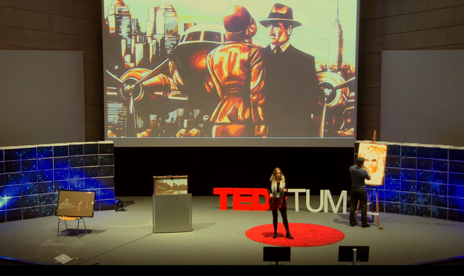 Audrey Sykes delivering TED Talk while Max Zorn creates tape artwork live on stage, with a large illuminated tape-art piece projected in the background.