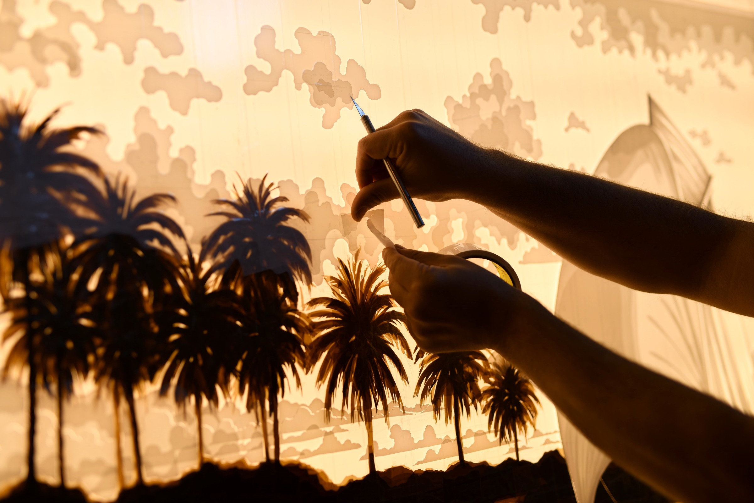 Max Zorn works with tape and a precision knife on Sail, with palm trees, ocean and a sailboat behind his hands.