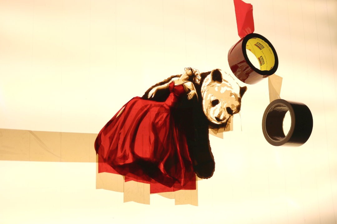The great escape in progress: a woman in a red dress beside a panda, with coloured tape rolls against the illuminated panel.